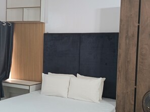 A bedroom with a bed, a nightstand, a wooden headboard, and a window with blinds. - Kbazzapartments (Ibadan)