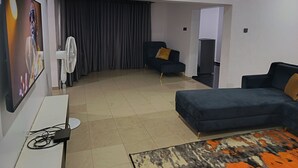 A living room with a sofa, a chair, a TV, a fan, and a patterned rug. - Kbazzapartments (Ibadan)