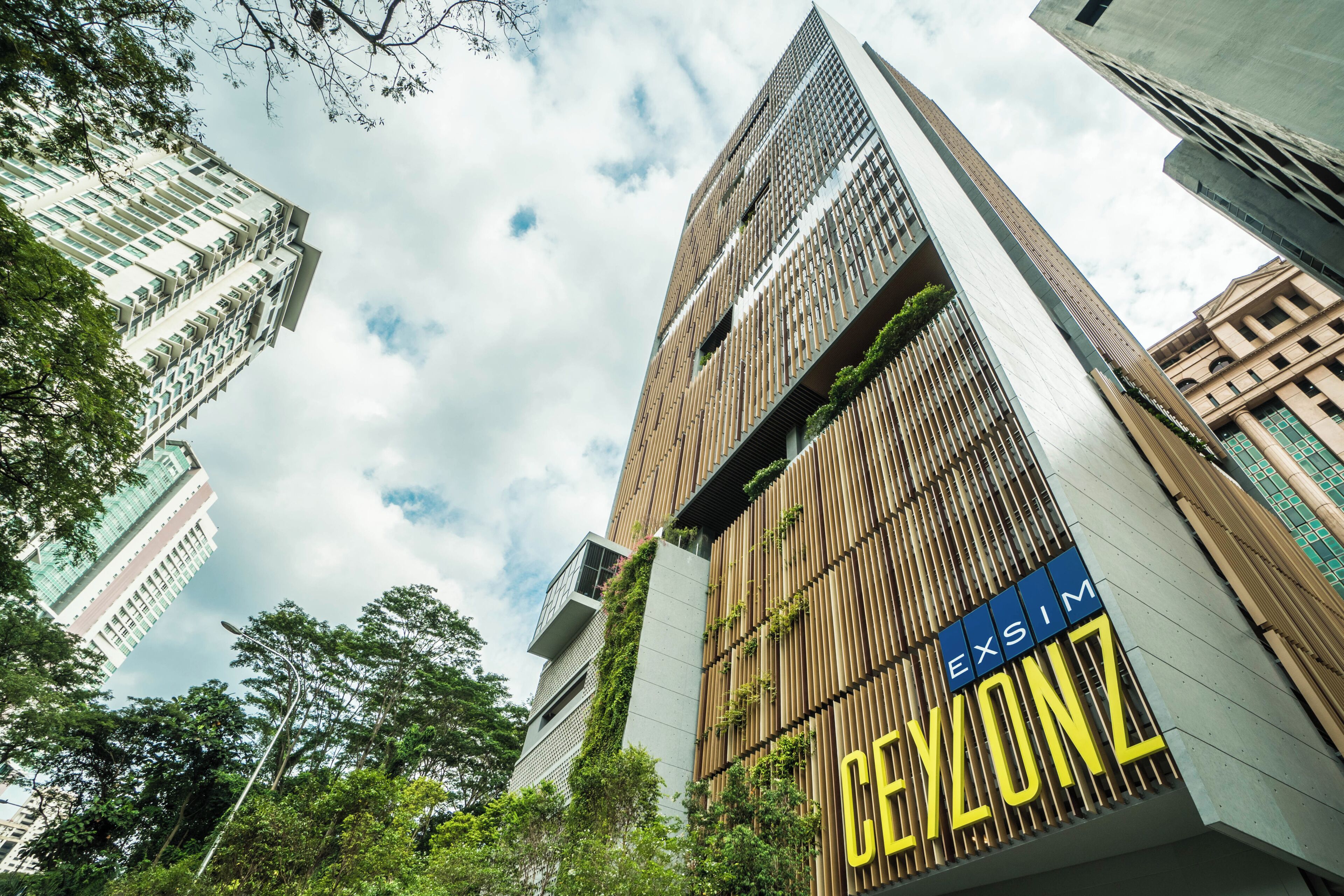 A modern building with a prominent 'EXIM CAYLONZ' sign, surrounded by other high-rise buildings and greenery.
