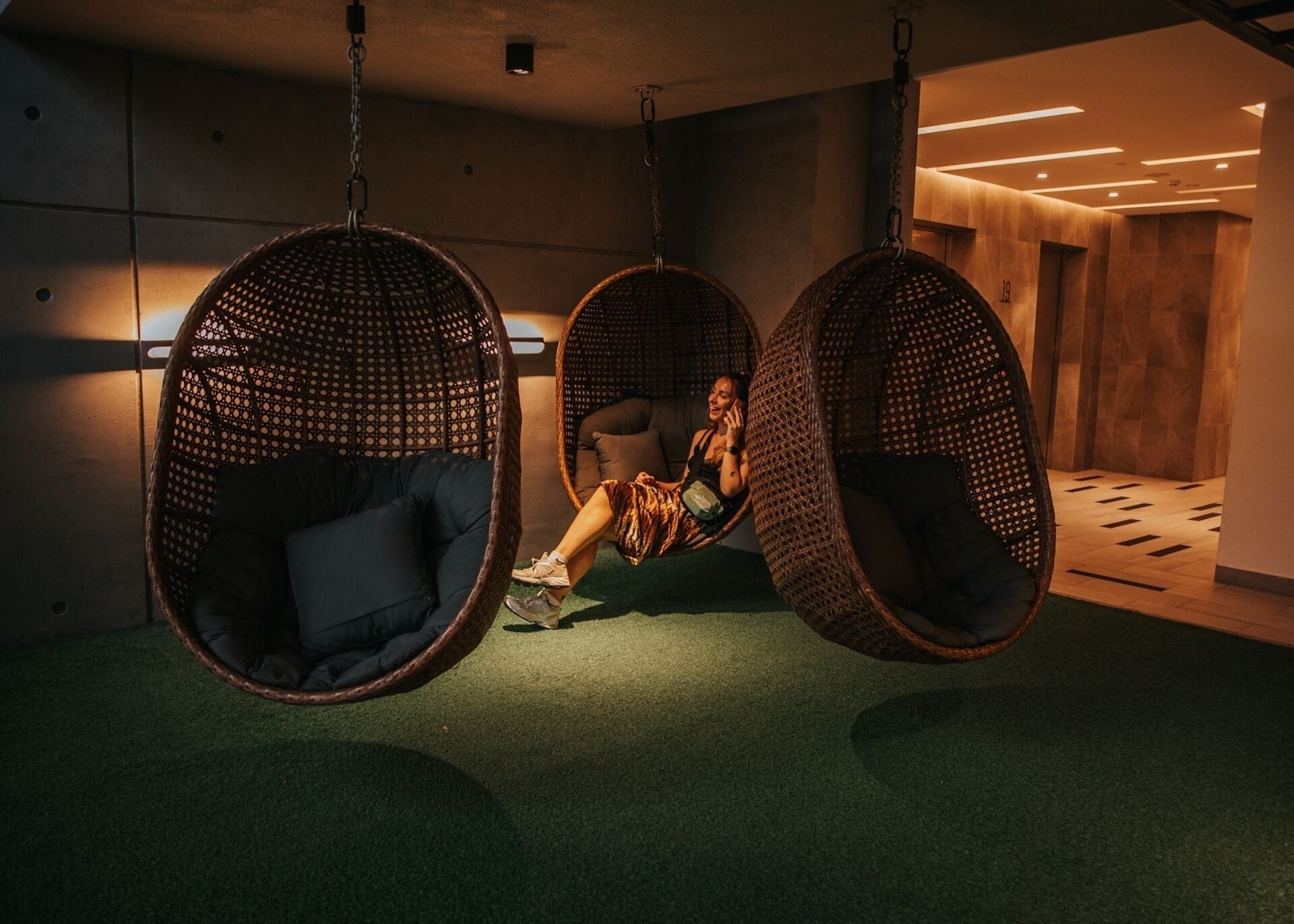 A modern interior with three hanging wicker chairs and a sofa, a green carpet, and a wooden floor.