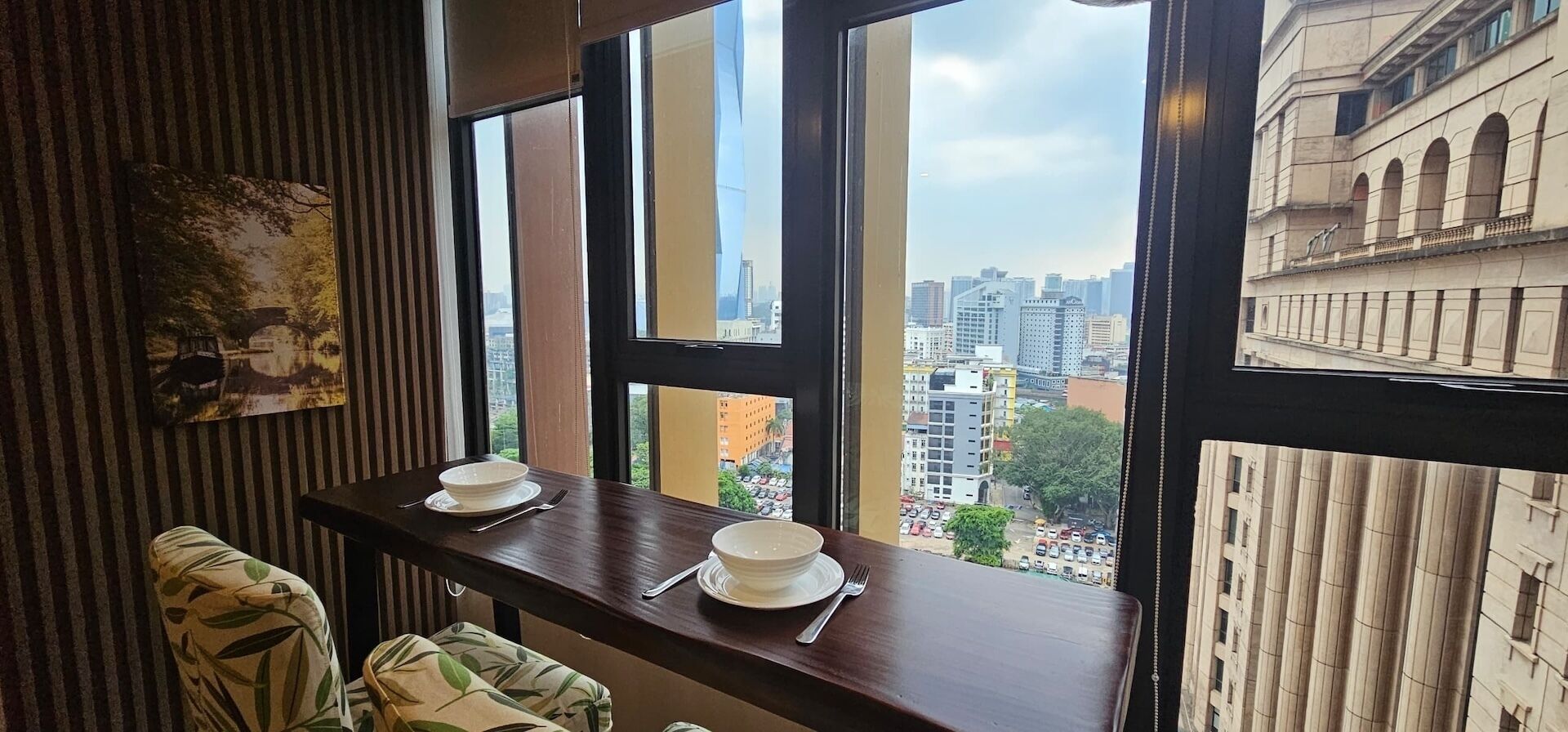 A dining area with a view of a cityscape through large windows, a wooden table, and a chair with a leaf-patterned cushion.