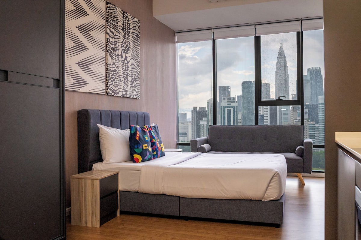 Un dormitorio moderno con una cama, un sofá gris, una mesita y vistas a la ciudad a través de grandes ventanales.