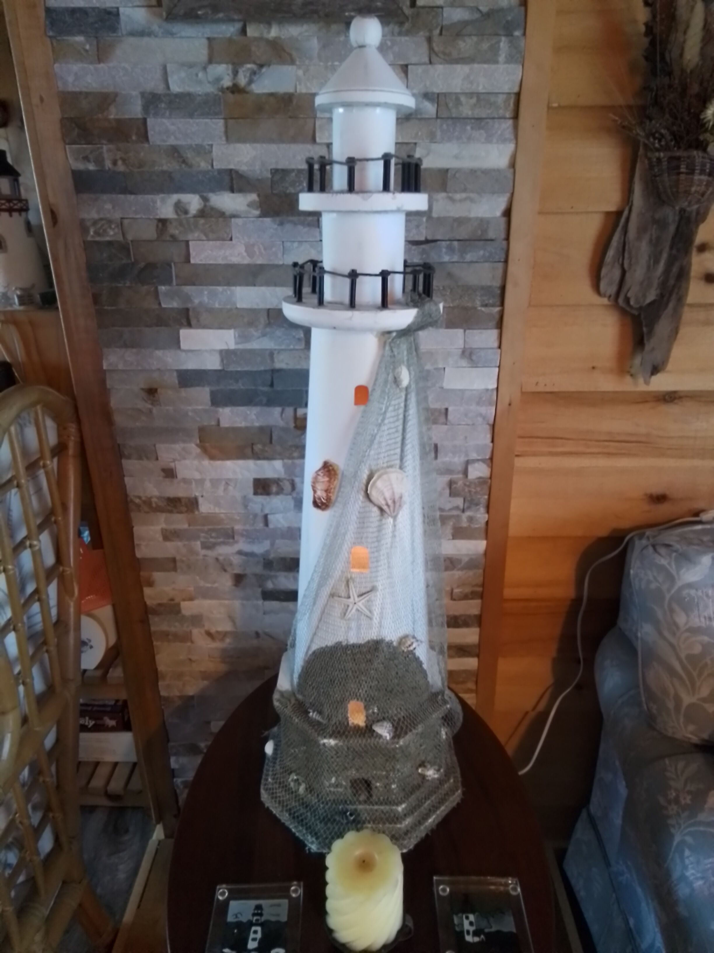 A lighthouse model with a net and shells, placed on a wooden table against a stone wall.