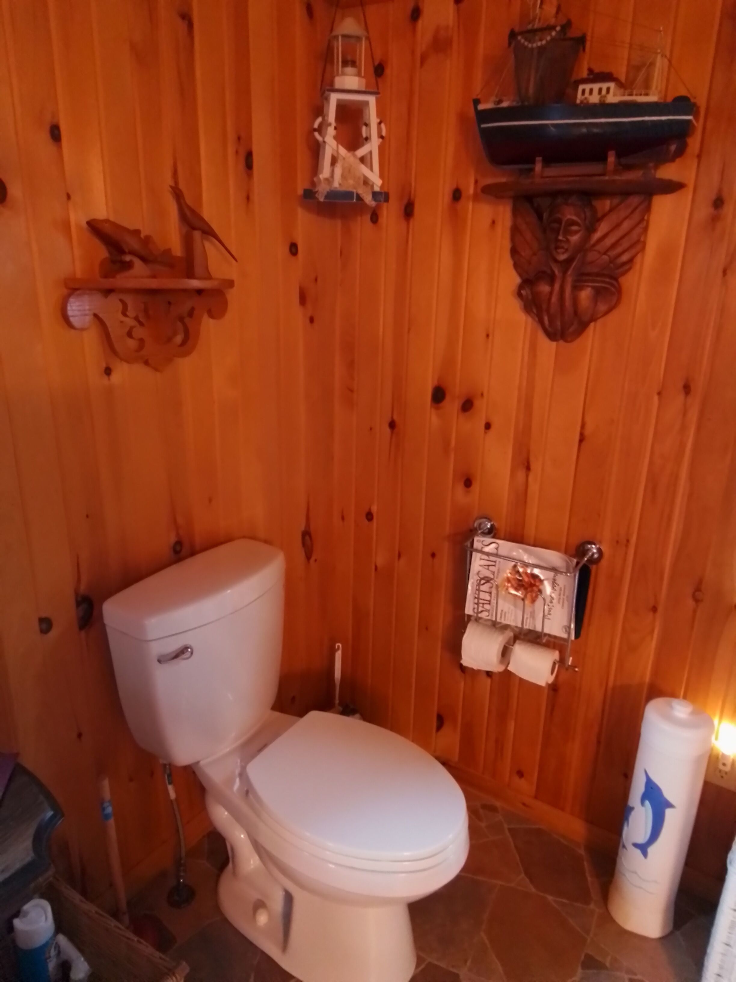 A wooden-paneled bathroom with a toilet, a hanging toilet paper holder, and a ship model on the wall.