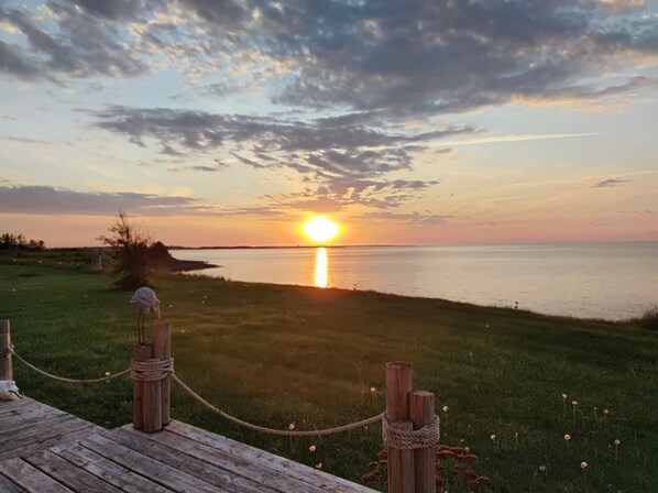 Un atardecer sobre una masa de agua, con una zona verde al frente y una terraza de madera.