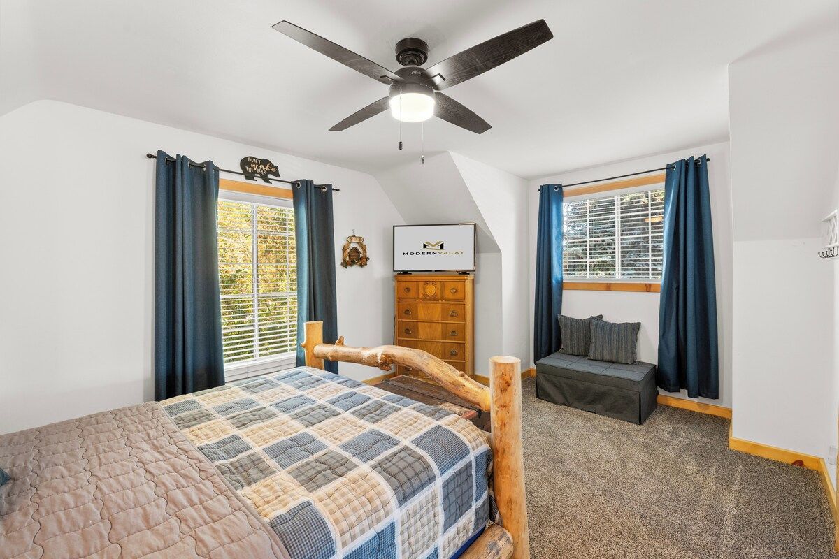 A bedroom with a bed, a wooden headboard, a ceiling fan, a dresser, a bench, and a window with curtains.