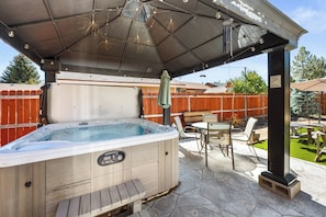 Ein Whirlpool unter einer überdachten Terrasse, mit Tisch und Stühlen, umgeben von einem Holzzaun und einer Naturstein-Terrasse.