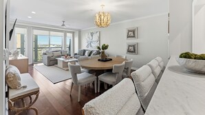 Ein modernes Wohnzimmer mit einem runden Esstisch, grauen Polsterstühlen, einem weißen Sofa und einem großen Fenster mit Blick.