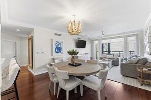 A modern living room with a round dining table, grey upholstered chairs, a chandelier, and a large window. - Residence 206 at The Grand (Wildwood)