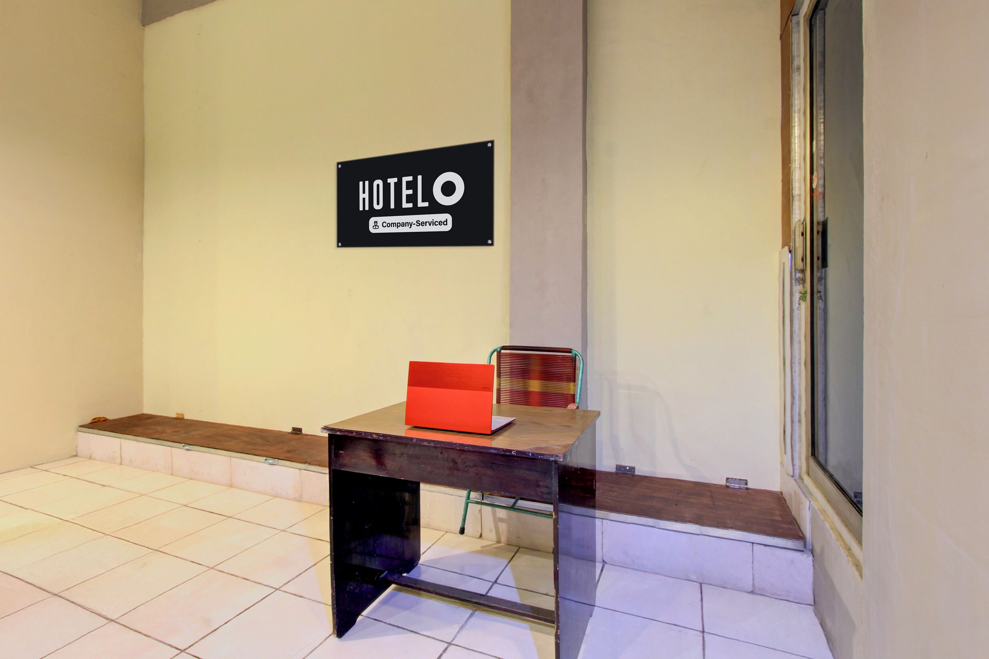 A hotel reception area with a sign indicating it is company-serviced, a desk with a red folder, and a window to the right.