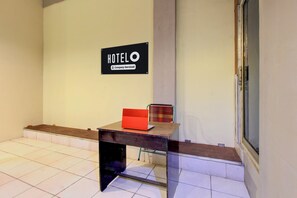 Un espace d’accueil d’hôtel avec un panneau indiquant qu’il est géré par l’entreprise, un bureau avec un dossier rouge et une fenêtre sur la droite.
