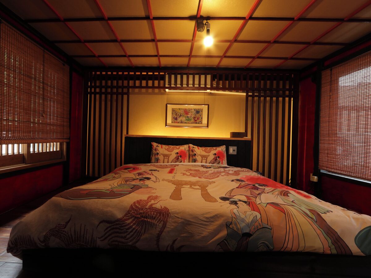 A traditional Japanese-style bedroom with tatami flooring, a low bed with a colorful bedspread, sliding shoji screens, and a ceiling with a visible light fixture.