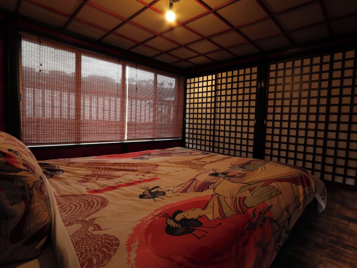 A traditional Japanese-style room with tatami flooring, a futon bed with a red and beige patterned bedding, sliding shoji screens, and a ceiling light fixture.