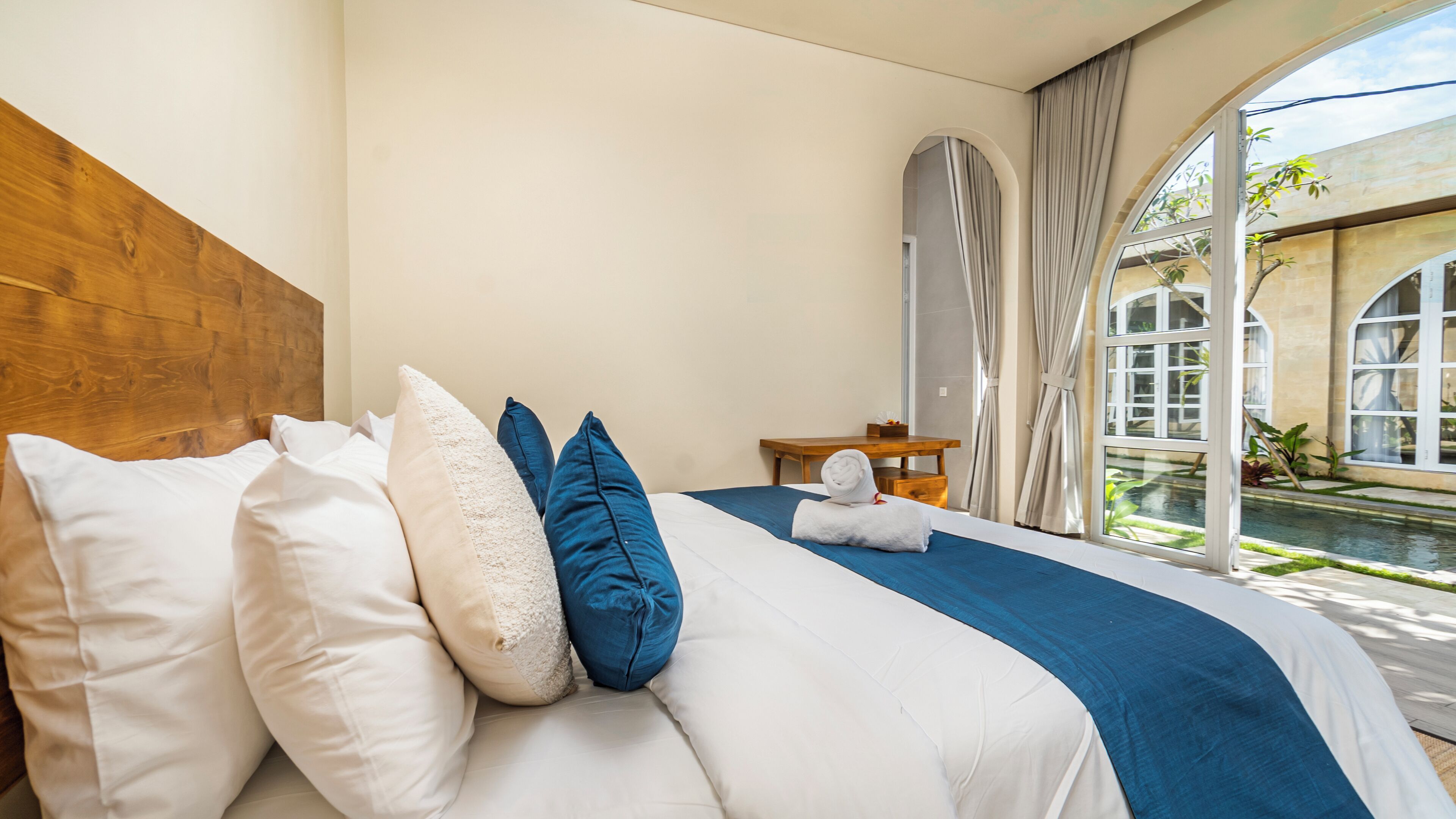 A hotel room with a bed, pillows, a wooden headboard, a nightstand, and a view of a courtyard with a pool.
