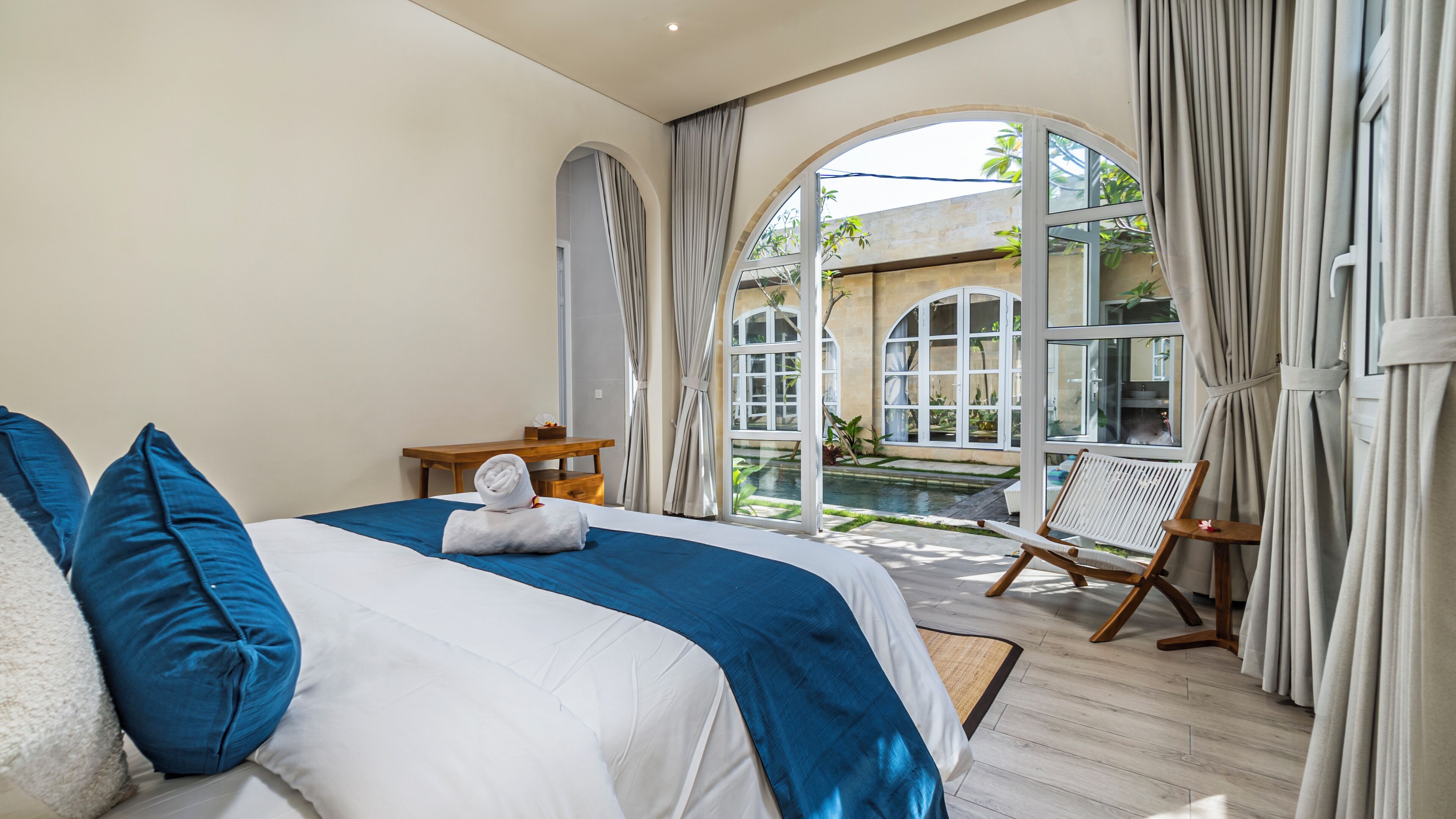 Une chambre dotée d’un grand lit, d’une table de chevet en bois, d’une chaise et offrant une vue sur une piscine et un jardin, grâce à de grandes fenêtres en arc de cercle.