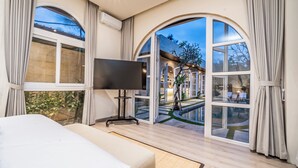 Habitación de hotel moderna con una cama grande, un televisor de pantalla plana y amplios ventanales de arco que ofrecen vistas del jardín y la piscina exteriores.