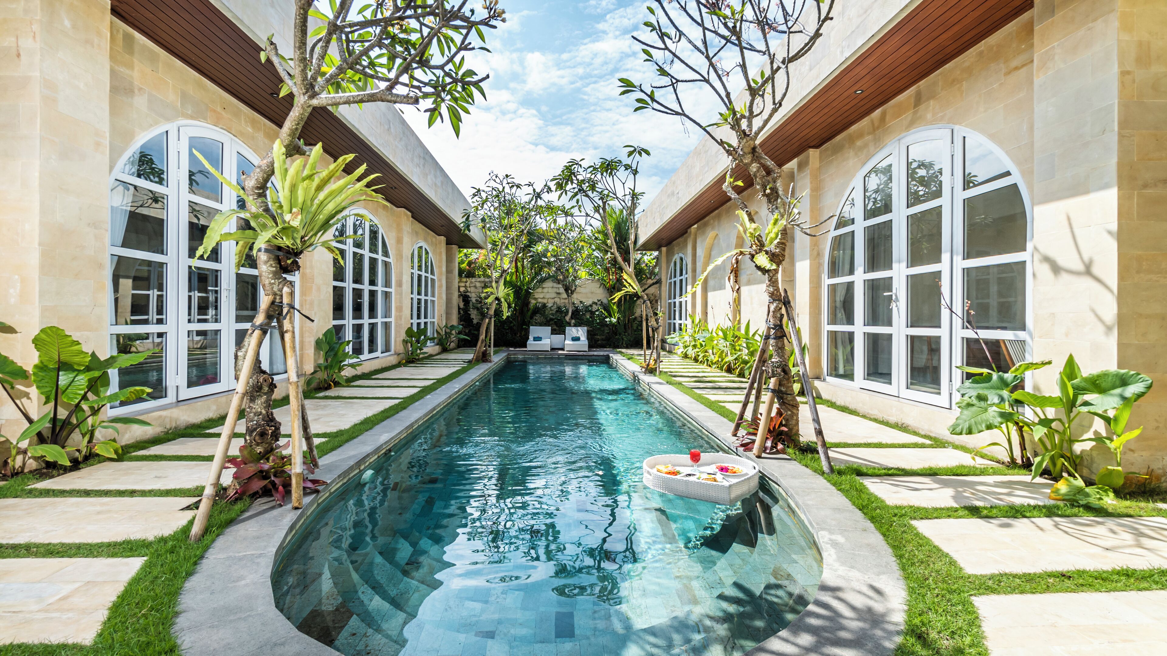 A swimming pool with a tiled deck, surrounded by lush greenery and two large arched windows on either side.
