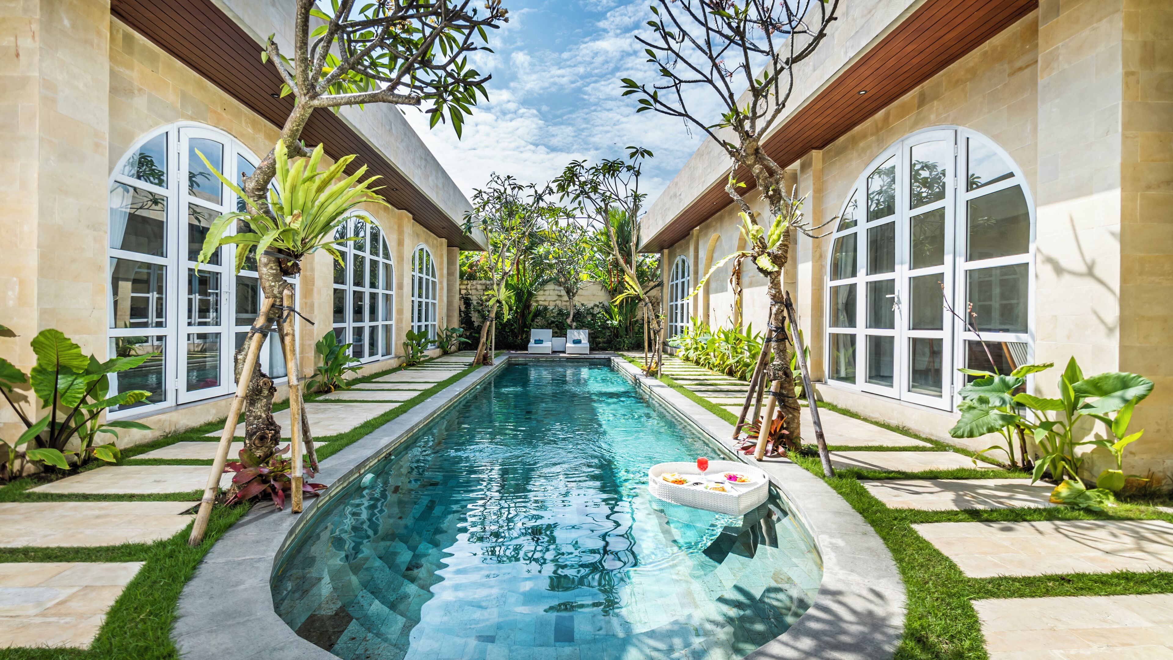 Ein Pool mit Fliesenrand, umgeben von einem Garten mit verschiedenen Pflanzen und Bäumen, und ein Gebäude mit großen Bogenfenstern an beiden Seiten.