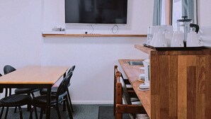 Um quarto com uma mesa de jantar de madeira, cadeiras pretas, uma televisão fixada na parede e uma janela com cortinas.