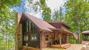 A rustic cabin with a stone chimney, wooden exterior, and a porch with an American flag. - Lazy Bear Cabin - Breathtaking Mountain Views, Hot Tub & Fire Pit (Clarkesville)