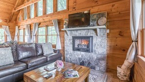 A log cabin interior with a stone fireplace, leather sofa, and a television mounted above it. - Lazy Bear Cabin - Breathtaking Mountain Views, Hot Tub & Fire Pit (Clarkesville)