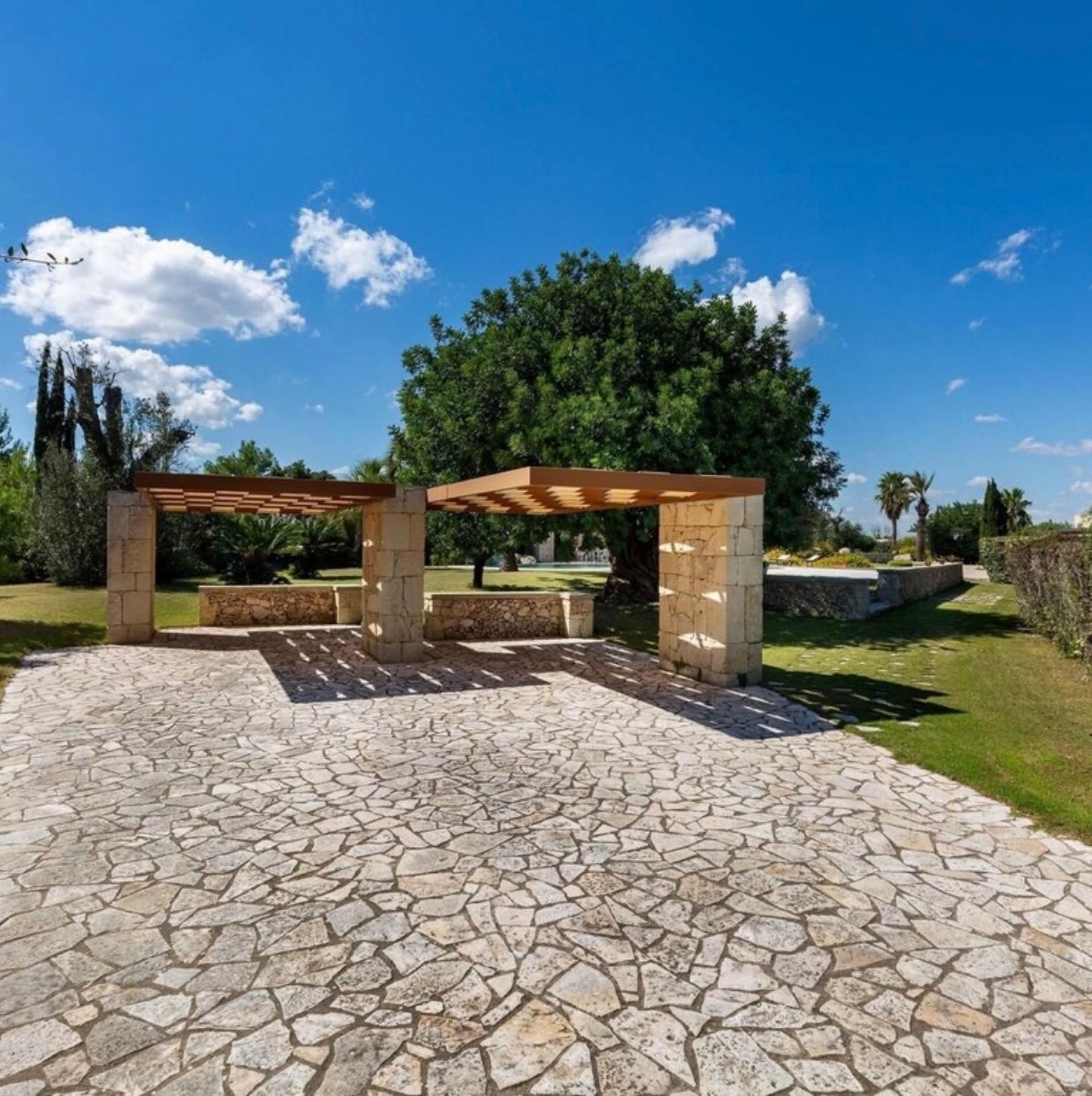 Un espace extérieur dallé de pierres, agrémenté d’une pergola et entouré de verdure et d’arbres.