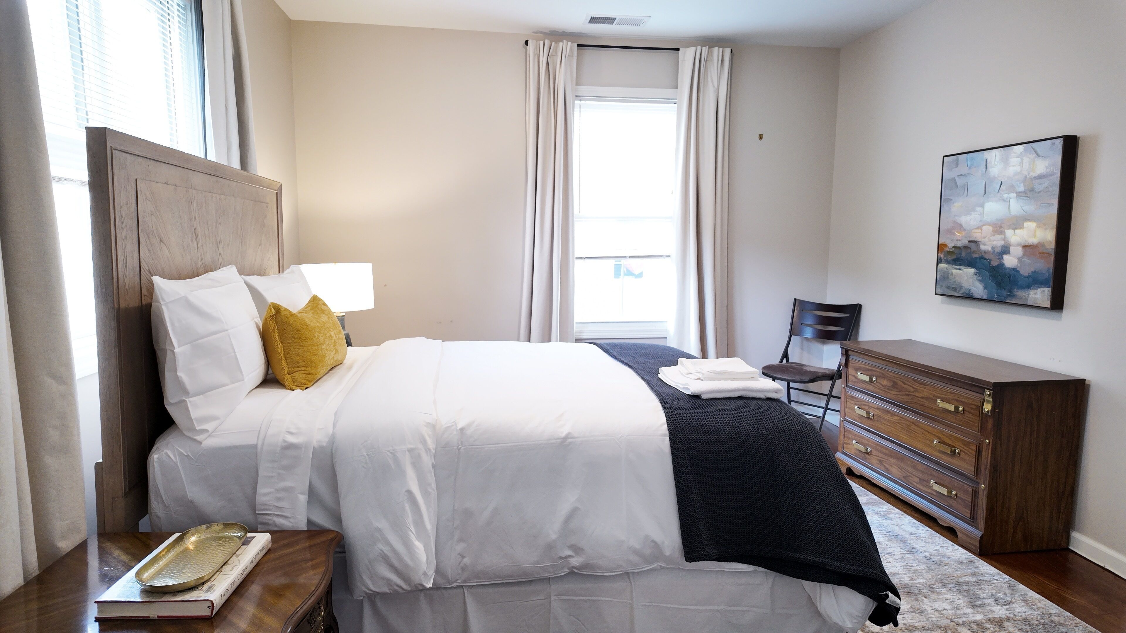 Quarto de hotel com cama, mesa de cabeceira, cômoda, cadeira, quadro e janela com cortinas.