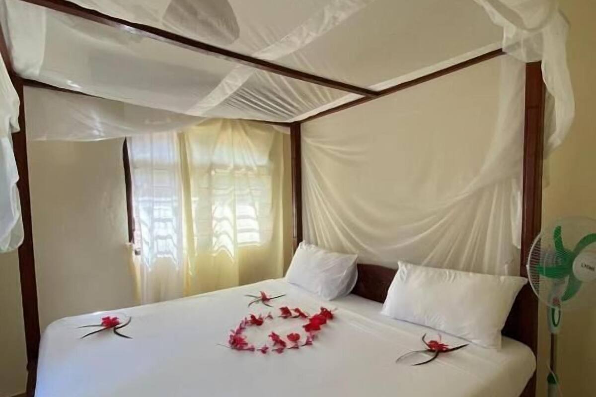 Un lit à baldaquin avec des draps blancs et un ciel de lit, un ventilateur sur la table de chevet et des fleurs rouges sur le lit.
