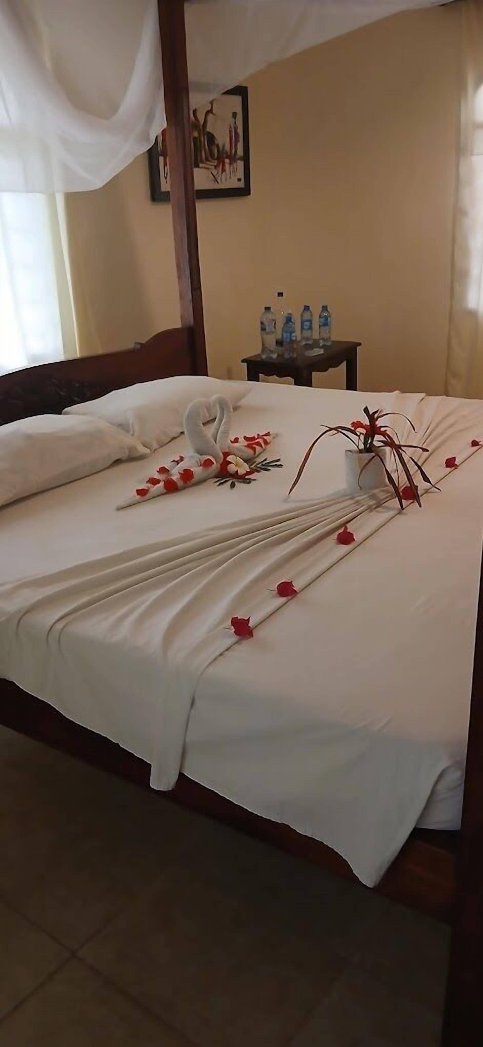A neatly made bed with white linens, a decorative swan pillow, and red rose petals.
