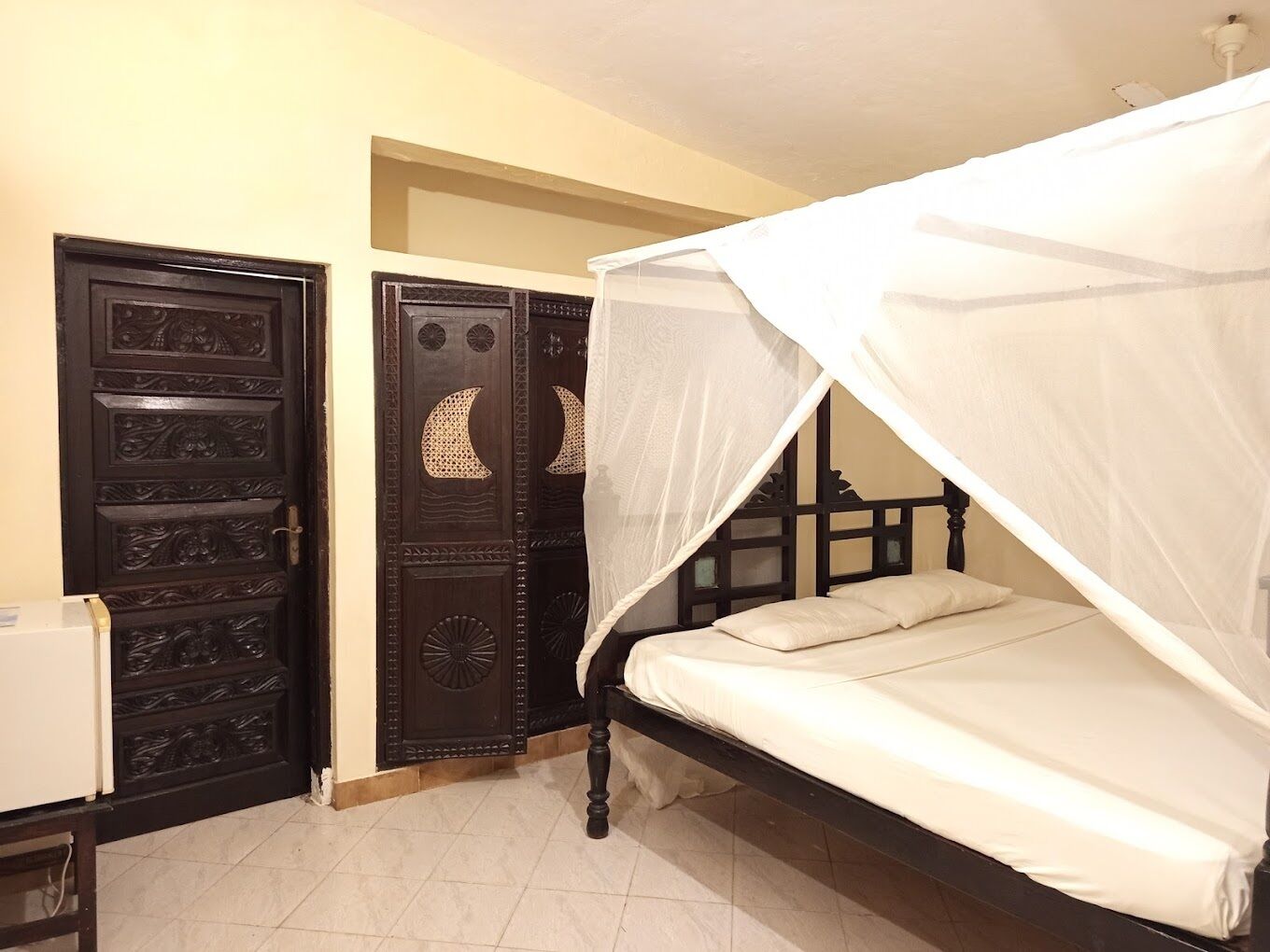 Quarto com cama sob um dossel, porta de madeira escura com painéis decorativos e parede branca.