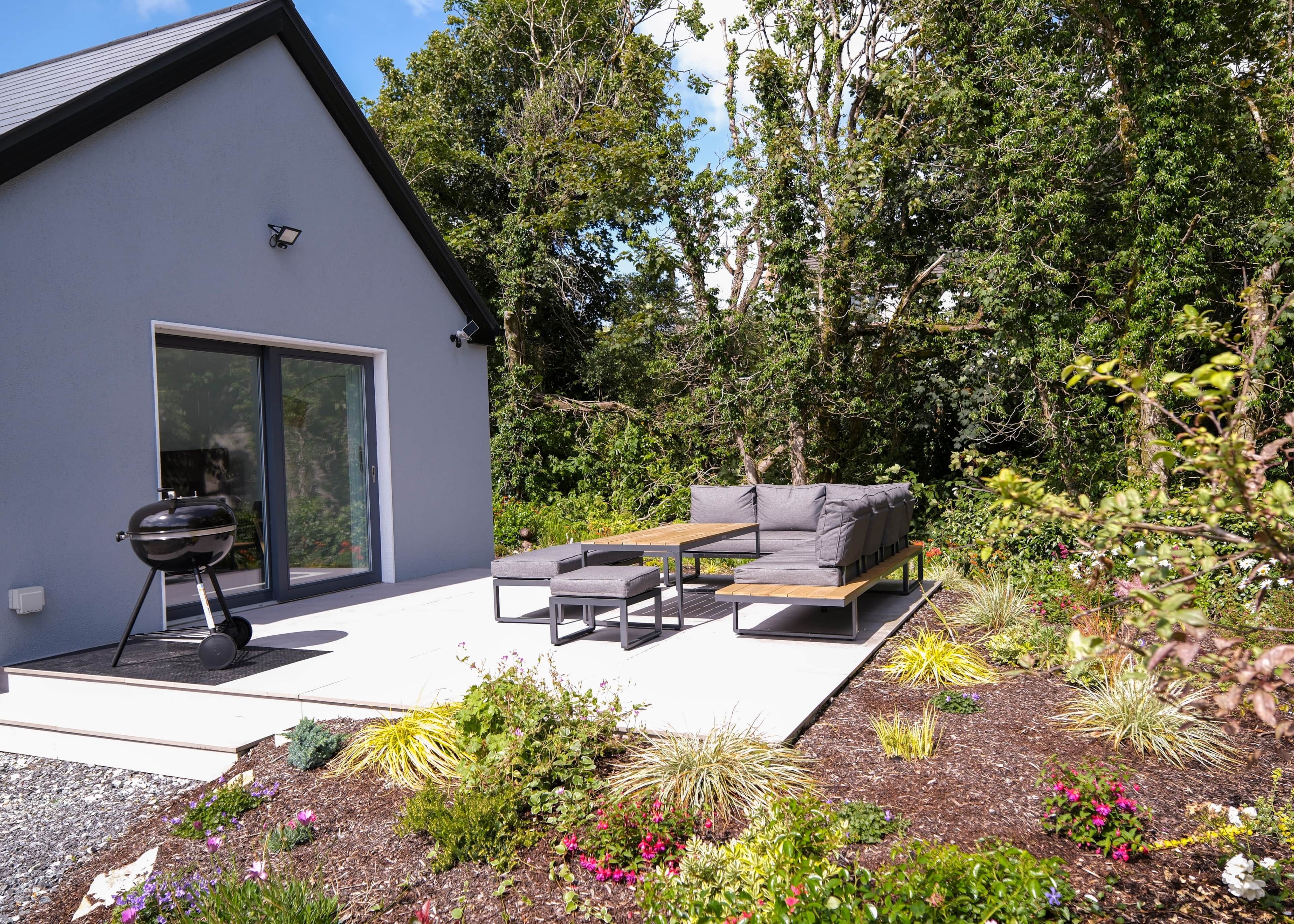 Un patio extérieur moderne doté d’un barbecue intégré, d’un canapé gris et d’une variété de plantes et de fleurs.