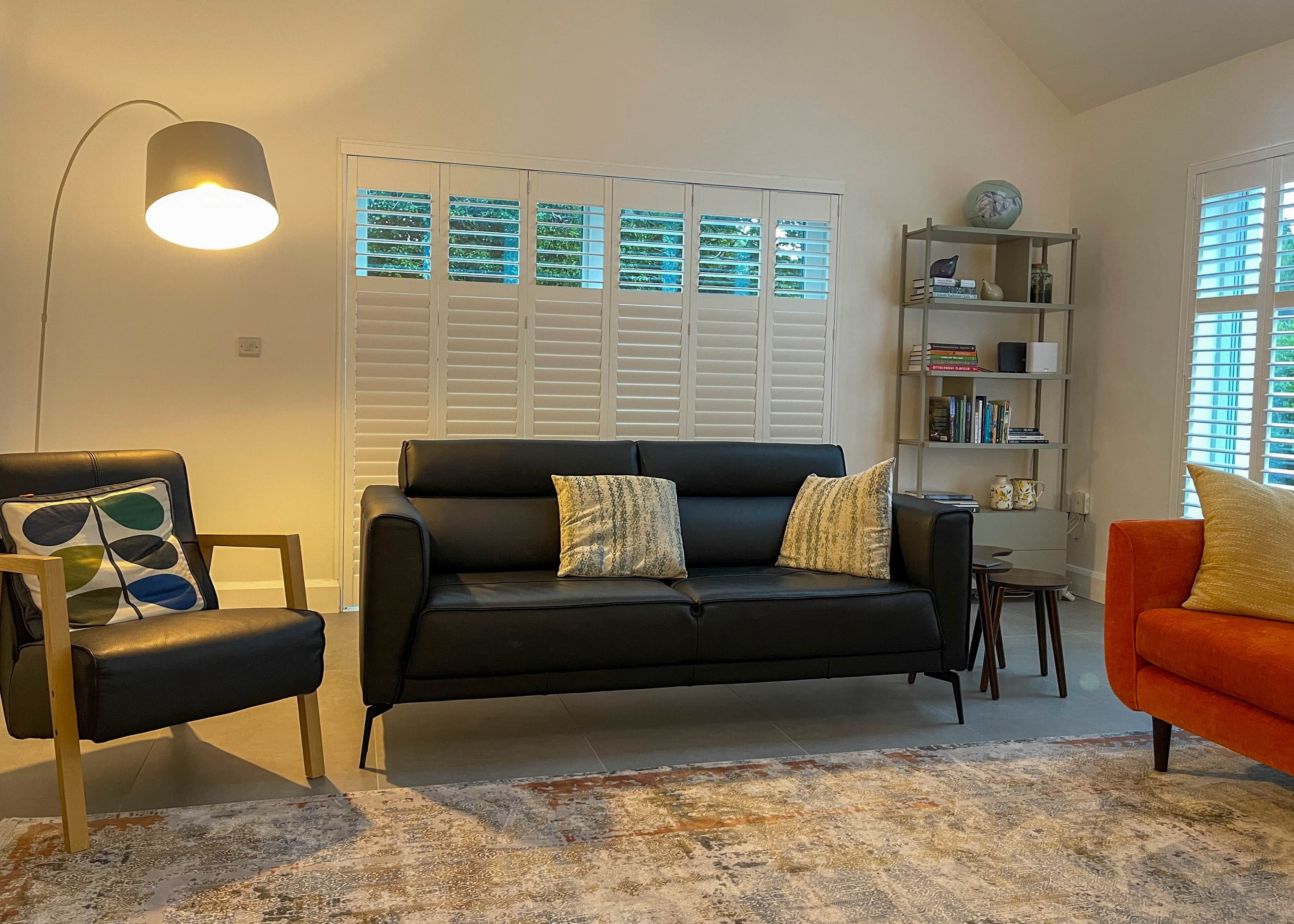A living room with a black leather sofa, a patterned rug, a bookshelf, and a window with blinds.