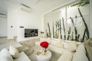 Un soggiorno moderno con una grande TV a schermo piatto, un divano beige, un tavolino da caffè con un vaso di fiori rossi e una vetrina con piante di cactus.