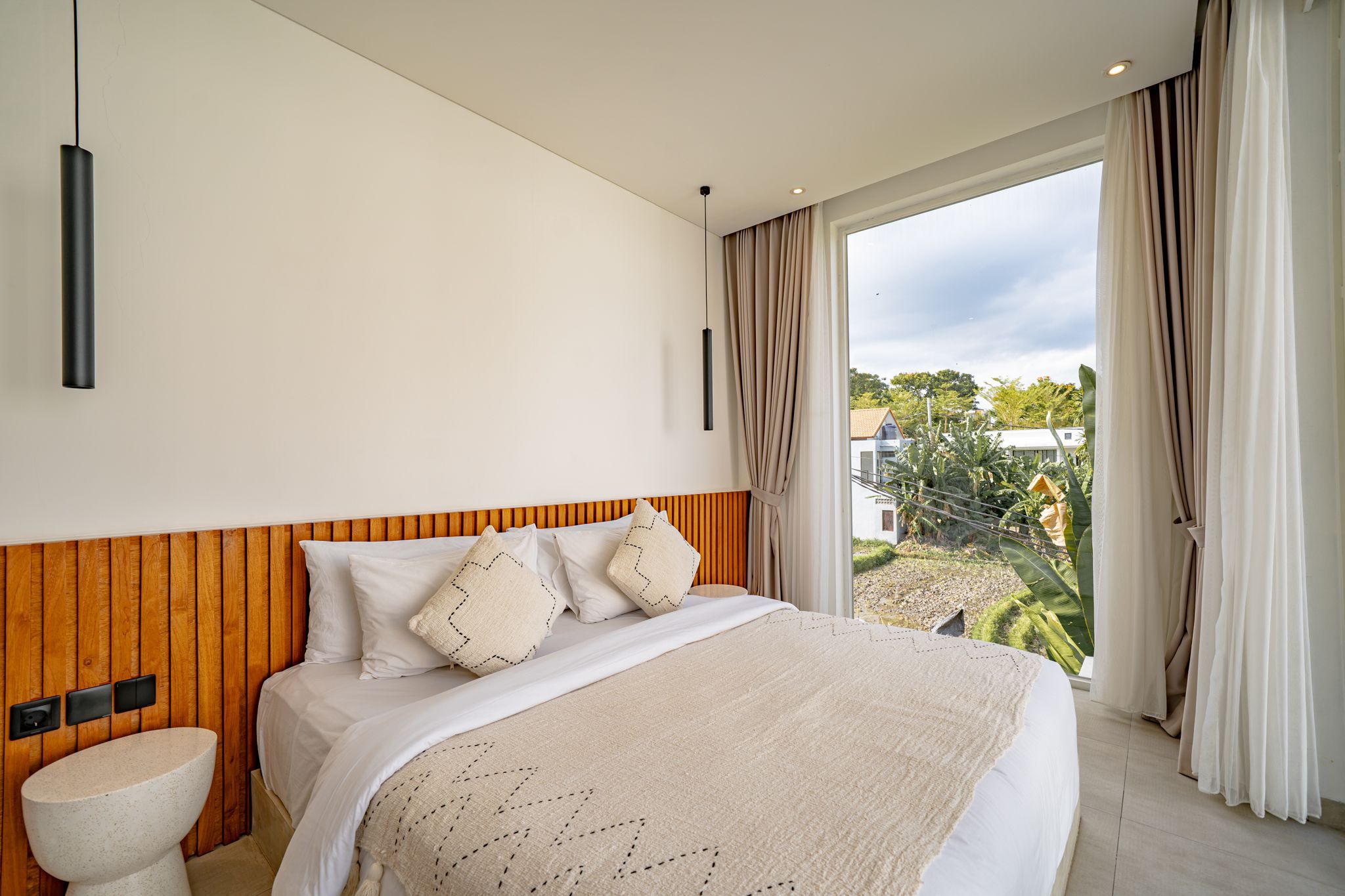 A modern bedroom with a large bed, a wooden headboard, a bedside table, and a view of the outdoors.