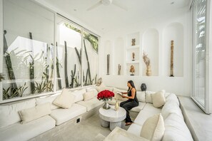 A modern living room with a white sofa, a coffee table, and a large window with plants. - The Belluoccio Villa (Canggu)