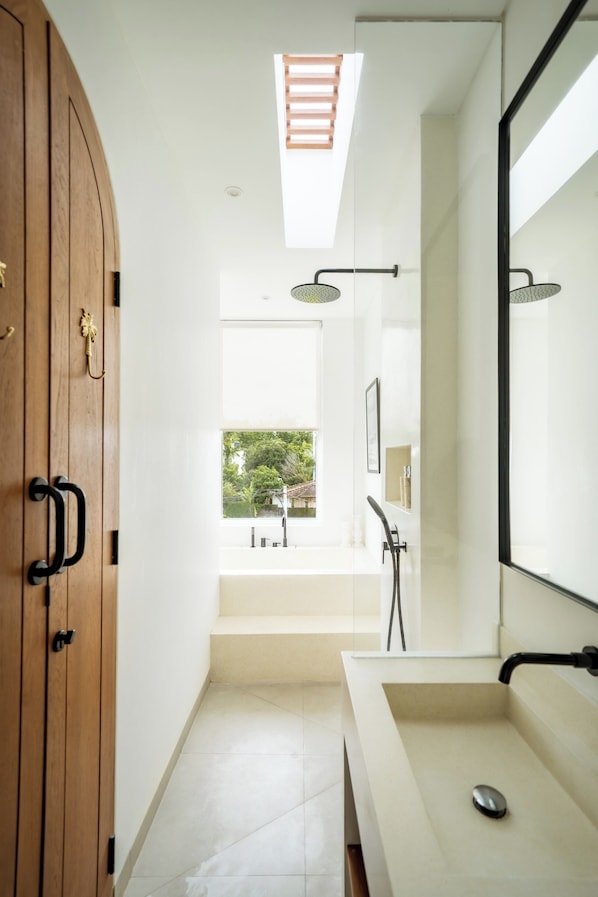 Un baño moderno con un espejo grande, una puerta de madera, un lavamanos blanco y un tragaluz.