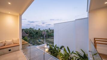Un balcón moderno con barandilla de vidrio, un sofá y vistas a un jardín y edificios al fondo.