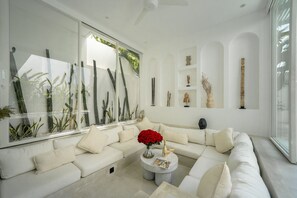 A modern living room with a white sofa, a coffee table with a vase of red flowers, and a large window with plants. - The Belluoccio Villa (Canggu)