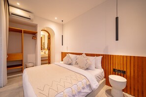 A modern bedroom with a bed, a wooden headboard, a bedside table with a round sink, and a walk-in closet with a mirror. - The Belluoccio Villa (Canggu)