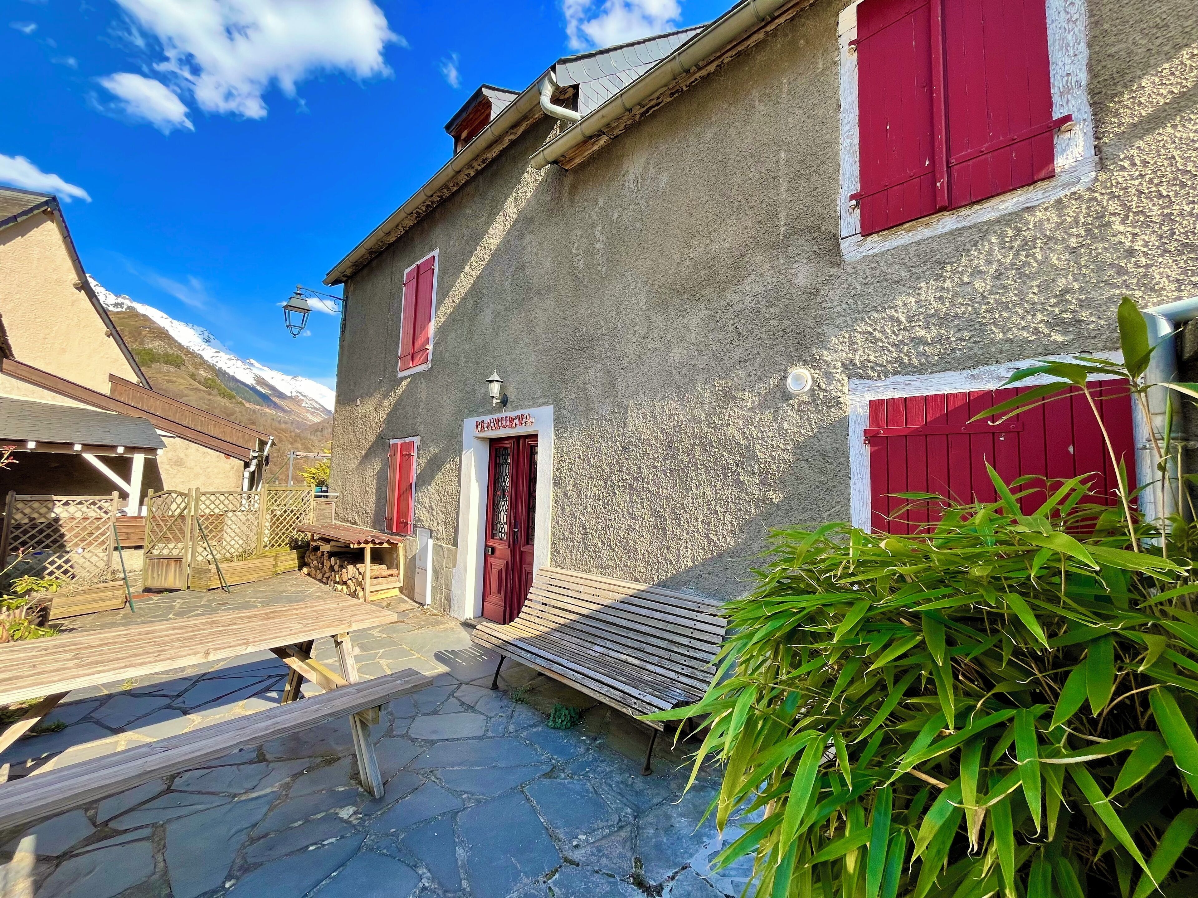 Un bâtiment rustique avec une façade en pierre, des volets rouges et un banc en bois au premier plan.