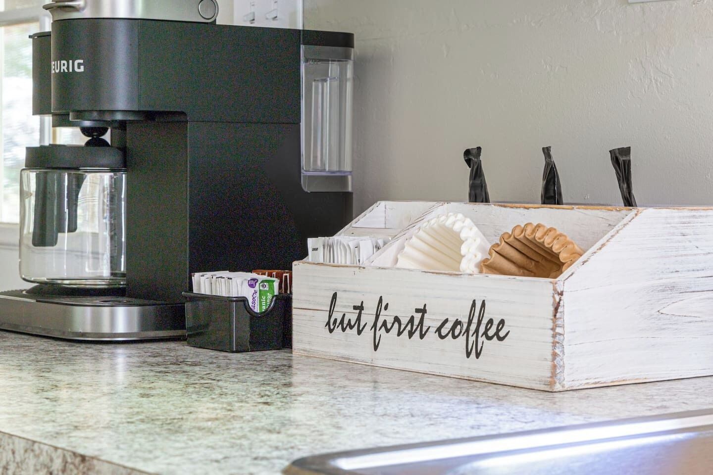 A coffee maker, a box labeled 'but first coffee,' and a coffee grinder on a countertop.