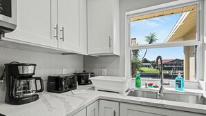 Une cuisine avec des meubles blancs, une machine à café, un grille-pain, un évier et une fenêtre donnant sur une maison et des palmiers.