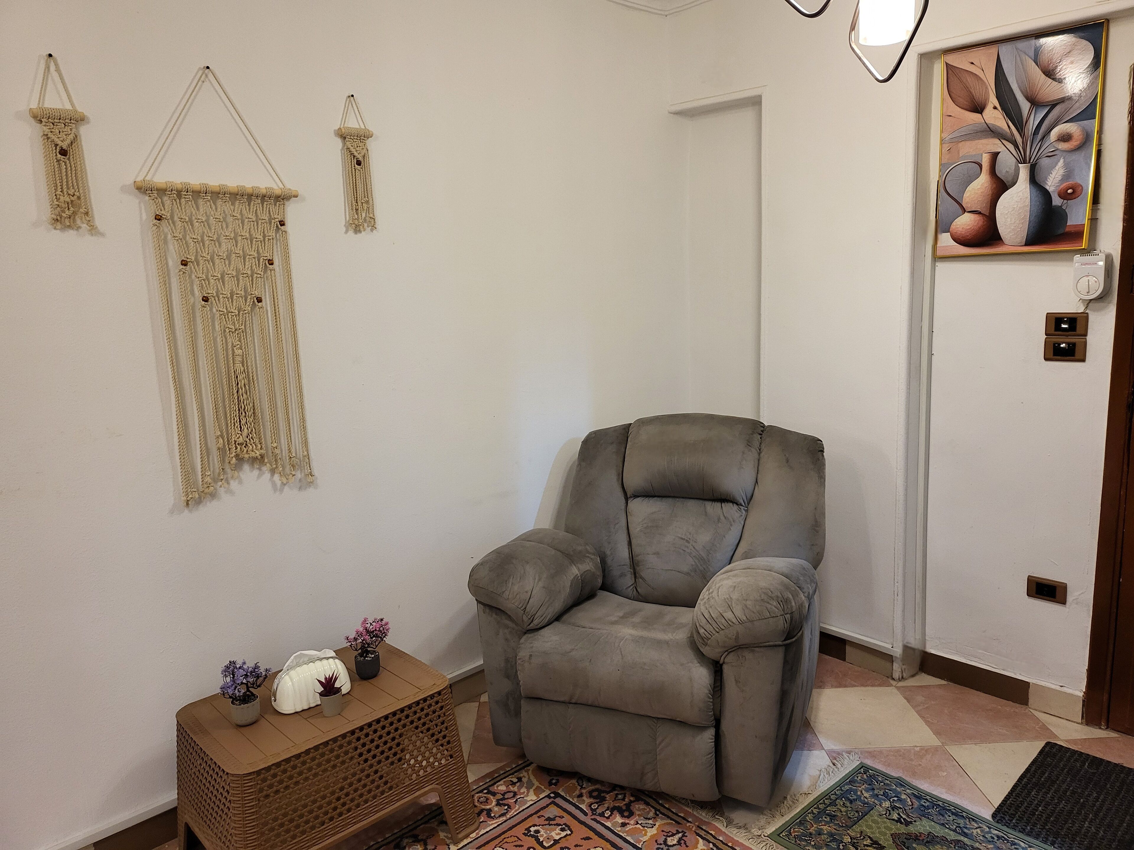 A living room with a grey armchair, a small wooden table with decorative items, macramé wall hangings, and a framed artwork on the wall.
