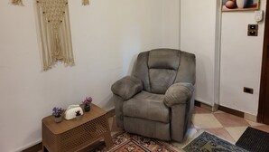 En stue med en grå lænestol, et lille træbord med dekorative genstande, makramovægge og et indrammet kunstværk på væggen.