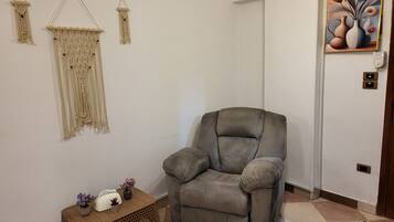 A living room with a grey armchair, a small wooden table with decorative items, macramé wall hangings, and a framed artwork on the wall.