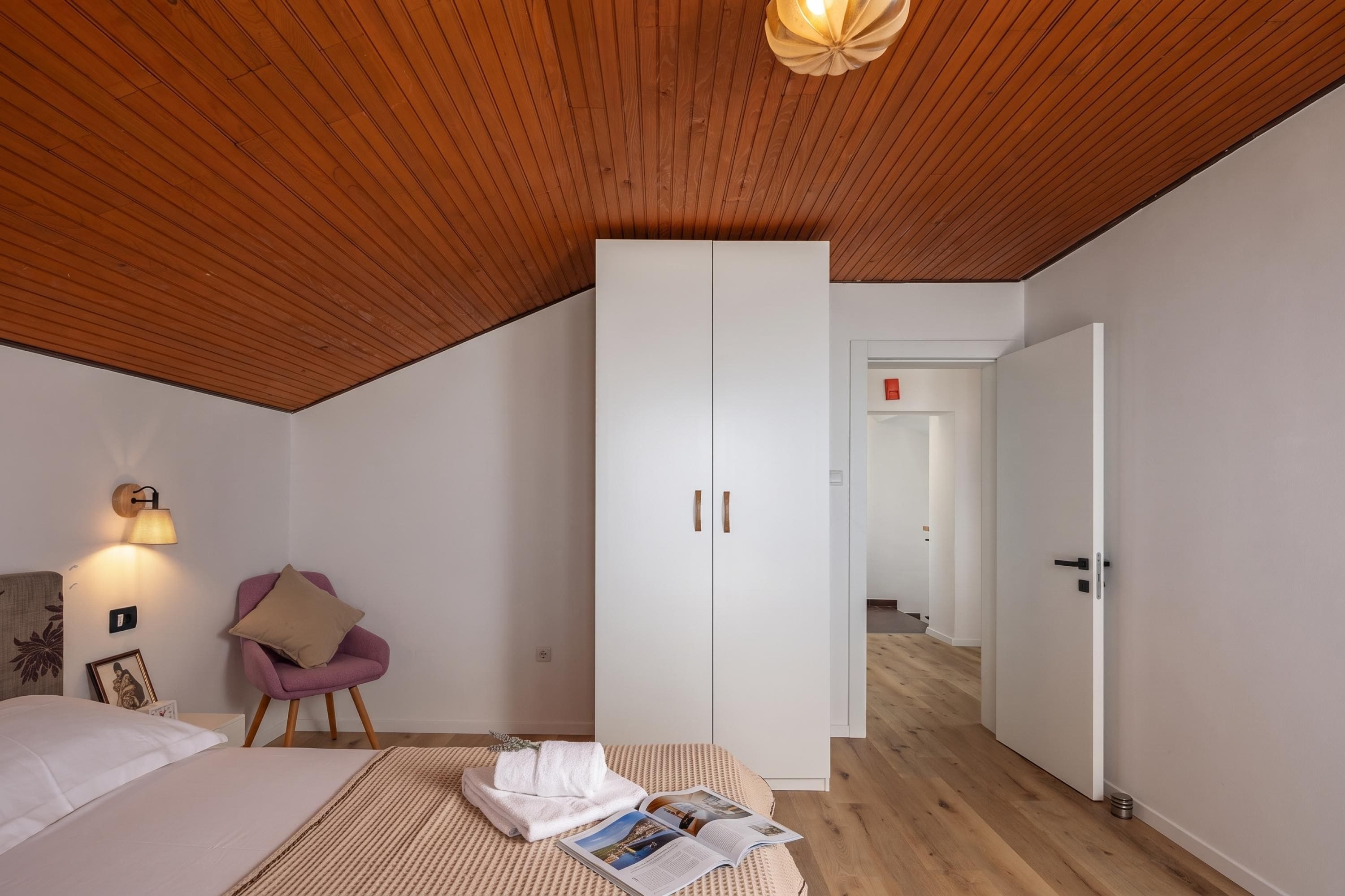 A bedroom with a sloped ceiling, a bed, a wardrobe, a chair, and a reading area with a magazine.