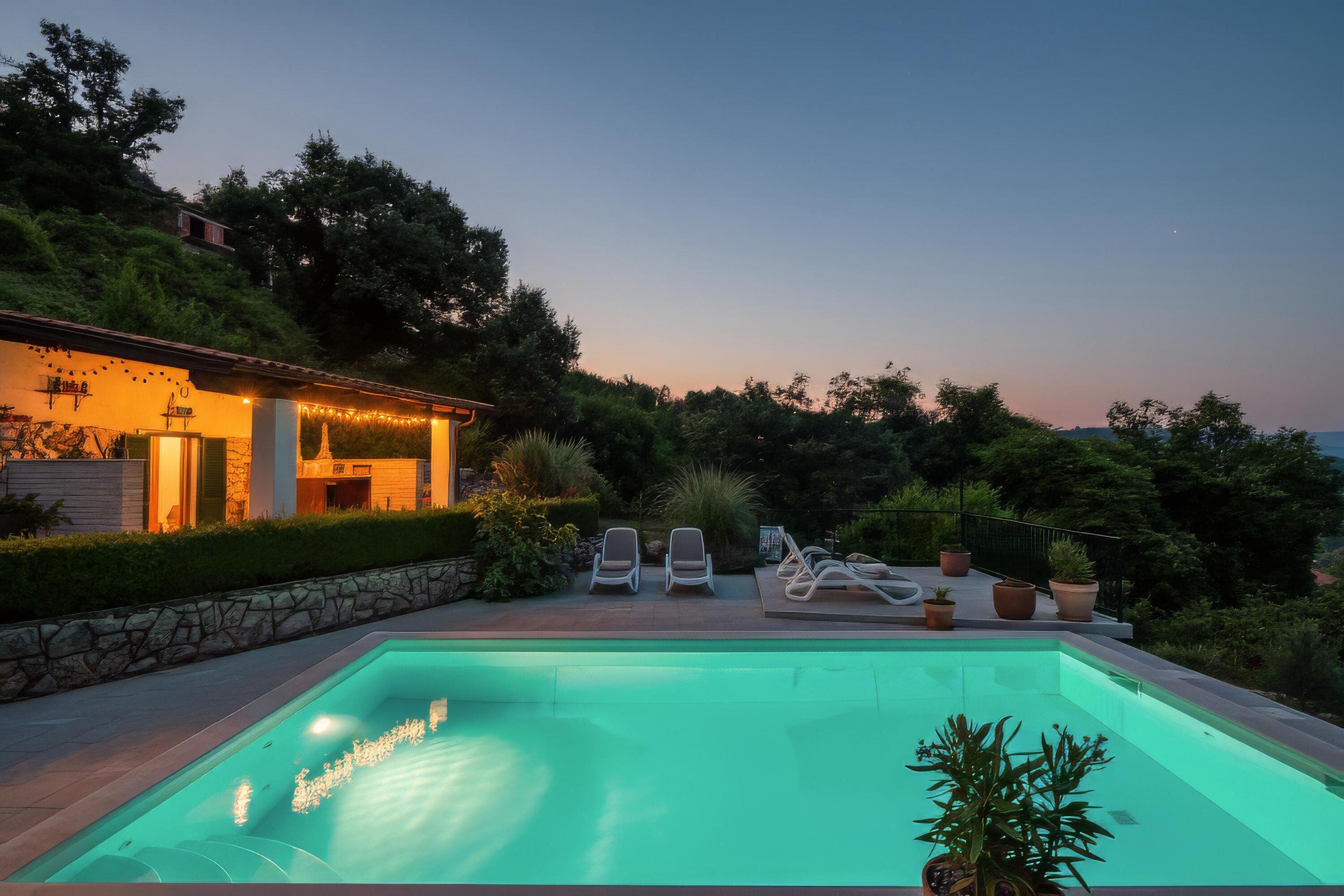 A swimming pool with lounge chairs, a stone wall, and a building with string lights.