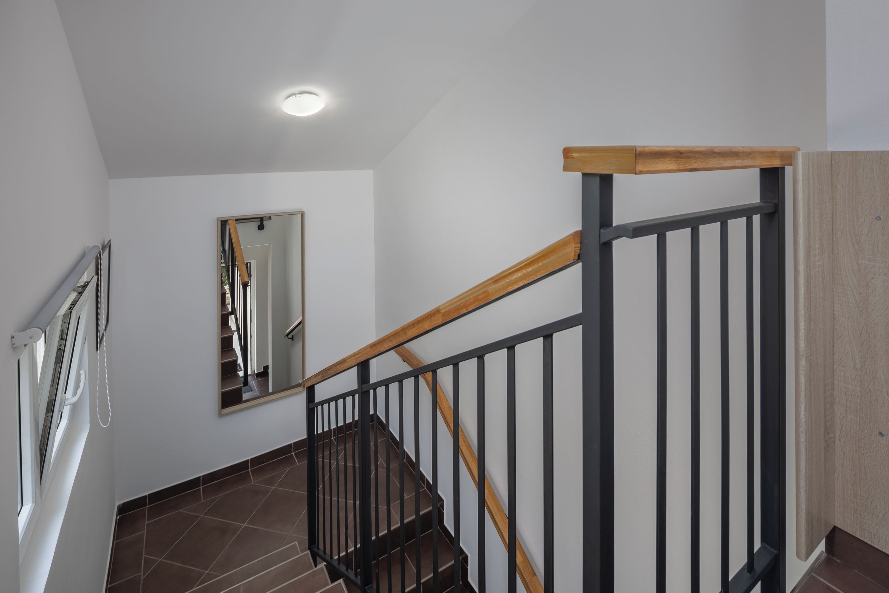 A staircase with wooden handrails and black metal balusters, a mirror on the wall, and a window with blinds.