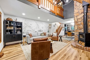 A spacious living area with a fireplace, a staircase, and a kitchen visible in the background. - Forest Hideaway with Hot Tub, Custom Fire-Pit, BBQ Deck & Cozy Mountain Vibes, Minutes to Mountain (Mount Hood Village)