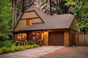 A two-story house with a garage, surrounded by dense forest. - Forest Hideaway with Hot Tub, Custom Fire-Pit, BBQ Deck & Cozy Mountain Vibes, Minutes to Mountain (Mount Hood Village)